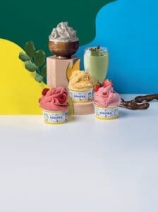 Colorful display of three gelato cups topped with fruit, set against a vibrant background of blue, green, and yellow. A wooden bowl of whipped cream and a glass of green smoothie are in the back, beside decorative leaves and branches.