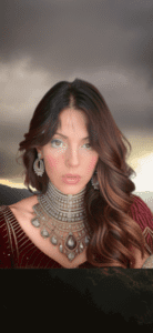 A woman with long, wavy hair is wearing an ornate necklace and earrings. She is dressed in a richly adorned red outfit. The background is a dramatic, cloudy sky with a hint of sunlight over silhouetted hills.