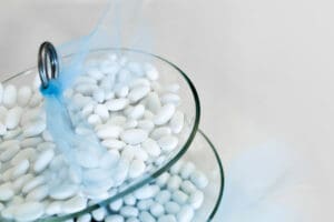 Two glass trays stacked, filled with white almond-shaped candies. A light blue mesh decorates the top tray, with a silver ring on the edge. The background is soft and light, enhancing the candies' sleek surface.