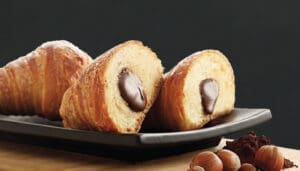 Croissants filled with chocolate cream are displayed on a black rectangular plate. The front croissant is cut in half, revealing the creamy filling. In the foreground, there are hazelnuts and cocoa powder on a wooden surface.