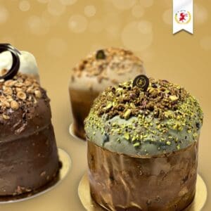 Three gourmet panettone cakes on a soft gold background. The nearest cake is topped with green frosting and pistachios. The others feature various nuts and chocolate. A small white ribbon with a red lion emblem is in the upper right corner.
