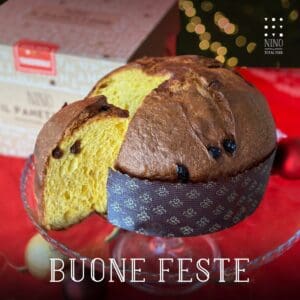 A golden-brown panettone with raisins, partially sliced, sits on a glass cake stand. It rests on a red cloth with holiday ornaments. In the background, there's a decorative box. Text reads "Buone Feste.